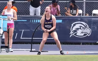 A young woman with a blond pony tail wearing a track singlet cheers while standing on the edge of the grass on a track.