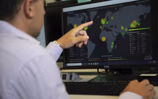 A man in a button-down shirt points at a computer displaying a world map and glowing with yellow and green circles. The map says "Cyber Attack Map Top Spam and Malware Senders" at the top.