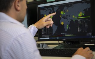 A man in a button-down shirt points at a computer displaying a world map and glowing with yellow and green circles. The map says "Cyber Attack Map Top Spam and Malware Senders" at the top.