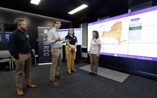 Governor Kathy Hochul stands with Nick Bassill and other New York emergency management leaders in front of a weather map inside the xCITE lab.
