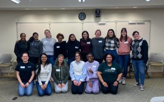 A group of 16 members of STEM NOW pose, all smiling, for a group photo in a classroom at UAlbany. The group is posed in front of a white board and they are arranged in two rows. The front row is kneeling on the carpet, which is gray. All are wearing white name tags.