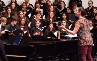 Young people dressed in all black are holding music books and singing on a stage with a piano and conductor.