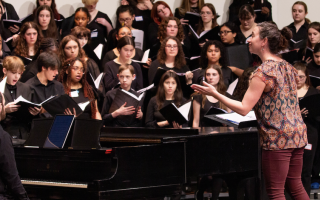 Young people dressed in all black are holding music books and singing on a stage with a piano and conductor.