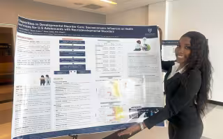 A young woman with long dark hair, wearing a black sweater and dress slacks, smiles as she displays her poster titled "Disparities in Developmental Disorder Care: Socioeconomic Influences on Health Services for U.S. Adolescents with Neurodevelopmental Disorders."