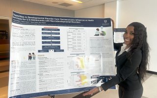A young woman with long dark hair, wearing a black sweater and dress slacks, smiles as she displays her poster titled "Disparities in Developmental Disorder Care: Socioeconomic Influences on Health Services for U.S. Adolescents with Neurodevelopmental Disorders."
