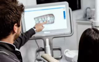 This image is of two people in a dentist office, taken from behind. A man with short brown hair points to a digital screen showing an image of a set of teeth. A person with long hair is seated behind him. 