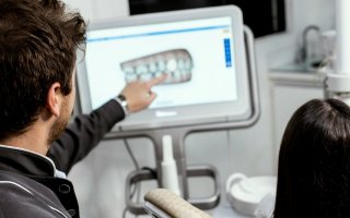 This image is of two people in a dentist office, taken from behind. A man with short brown hair points to a digital screen showing an image of a set of teeth. A person with long hair is seated behind him. 