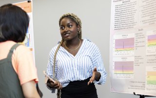 An African -American young woman with a white and blue vertical-striped blouse, a gold necklace and a black skirt gestures with her hands while speaking in front of an academic poster indoors.