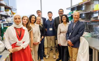 Nine smiling people stand together in a lab, posing for a group portrait. They are flanked by lab benches and shelving stocked with lab supplies. 