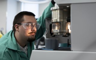 A man wearing glasses, protective goggles and a green lab coat handles a lab instrument that features a metal cylinder containing a lit glass chamber. 