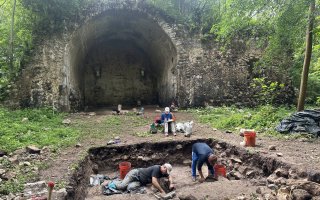 Archaeologists work in a hole digging with tools, with one person recording notes, in the background is an archway of a ruined church from the 1500s.