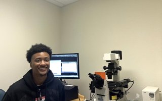 A smiling young man wearing a black jacket and clip on name badge sits in front of a computer screen next to a microscope.