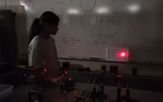 A girl with dark hair and a longsleeve shirt stands in front of a whiteboard with equations while a Raman spectroscopy laser is pointed at the wall behind her.