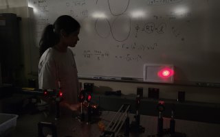 A girl with dark hair and a longsleeve shirt stands in front of a whiteboard with equations while a Raman spectroscopy laser is pointed at the wall behind her.