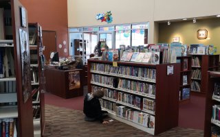 A boy looks at a series of books on a bookshelf while seated on the floor in a library.