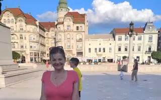 A woman in a pink blouse stands in front of a classical Roman large building in a city plaza with a blue sky and clouds in the background.
