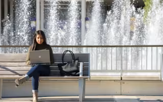 Student on laptop sitting on a bench in front of fountain