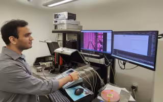 A man with short dark hair in a gray shirt stands in front of two computer screens in a laboratory.