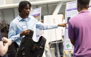 A man with short black braids in a light blue shirt holds up a black vest and gestures to a poster as he speaks to a man standing nearby in a purple shirt