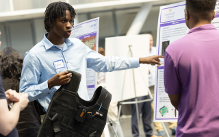 A man with short black braids in a light blue shirt holds up a black vest and gestures to a poster as he speaks to a man standing nearby in a purple shirt