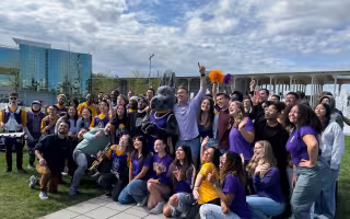 A crowd of people dressed in mostly purple, surround a person dressed in a Great Dane mascot outfit and a man with his hand raised, signaling number one, outside in a plaza against a blue sky with cloud cover.