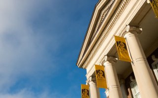A bright blue sky accentuates the roof of a brick building with ornate columns holding yellow UAlbany flags