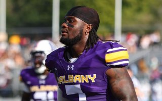 An African American man wears a bright purple jersey with the letters UALBANY across the front and the number 7. There is a football field, fans and other players in the background.
