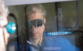 A image showing the reflection of a man in a blue lab coat and wearing blue safety goggles.