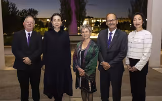 A group of five people stand in a row for a portrait, a man in a suit and purple tie, a woman in a long black coat, a shorter woman in a colorful shawl, a man in a gray suit and purple tie with a mustache and a woman in a white sweater or blazer against a night/dusk skyline with lights in the distance.