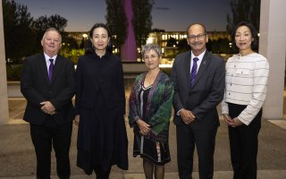 A group of five people stand in a row for a portrait, a man in a suit and purple tie, a woman in a long black coat, a shorter woman in a colorful shawl, a man in a gray suit and purple tie with a mustache and a woman in a white sweater or blazer against a night/dusk skyline with lights in the distance.