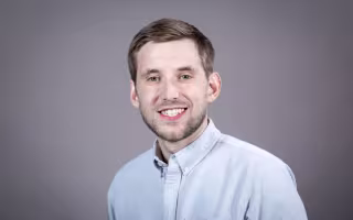 A man wearing a light blue button-down shirt smiles for a portrait against a gray backdrop.