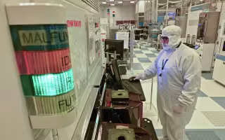 A man works at a computer terminal inside of a high tech clean room wearing a full white cleanroom suit, including white gloves and a white cap.