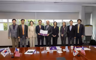 Nine people in business suits stand in front of a table topped with the American and South Korean flags and pose for a photograph