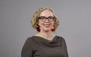 A profile photo of a woman with blonde hair, large red earrings and glasses wearing a dark brown blouse against a gray background.
