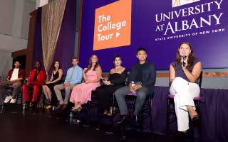 A group of students in colorful outfits sit on high chairs holding microphones in a line as part of a panel discussion, as a display behind them reads "The College Tour" and "University at Albany".