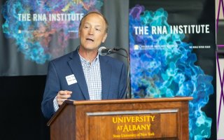 A man speaking at brown wood lectern with the words "University at Albany" written on the front in gold, in front of a colorful banner that says "The RNA Institute."