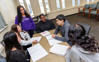 A group of students sit around a table with notebooks on a the table, one student. stands at the end of the table with a sweater that says Department of Languages, Literatures and Culture.