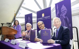 Four people are pictured under a white event tent with strong sunshine coming through. A woman wearing a purple blazer stands at a wooden podium. Three men wearing blazers sit at a table with a purple tablecloth, all smiling at the camera. Purple “University at Albany” banners are behind them.