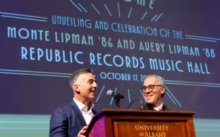 Avery Lipman (left) and Monte Lipman (right) speak at lecturn behind sign at the unveiling of the music hall named in their honor
