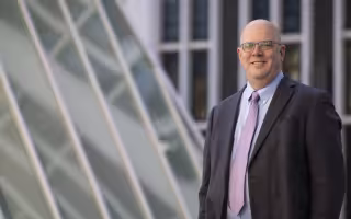 A smiling man in a gray jacket and purple tie stands outdoors near windows