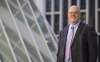 A smiling man in a gray jacket and purple tie stands outdoors near windows