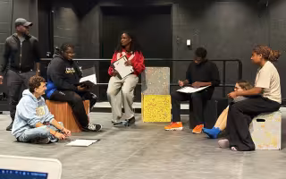 a group of theatre students and faculty, one standing to the left, the rest seated on the floor or boxes, look over scripts and notes in a flat theatre space with dark or black walls behind them.
