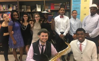 A group of people in 1920s attire pose for a photo. One is holding a saxophone and another is holding The Great Gatsby book.