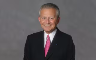 A man with silver hair in a black suit and pink tie smiles for a portrait against a gray backdrop.
