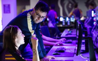 A man in glasses leans over one of a bank of computers, speaking to a younger man with long brown hair.