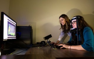 A woman in an apparatus looks at a monitor measuring where her line of sight goes as anothe woman looks on.