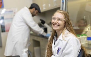 A woman with long wavy blond hair, wearing a white lab coat and clear protective goggles, smiles for a portrait in a lab. Lab benches with test tubes, and a man looking into a microscope, are in the background.