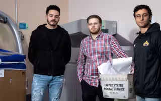 Three students stand indoors in front of a shredding machine and bypass that they created.