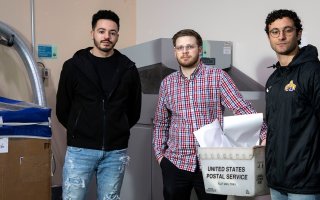 Three students stand indoors in front of a shredding machine and bypass that they created.