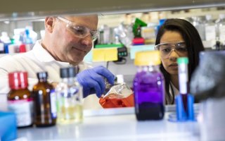 A photo of several glass bottles filled with colorful liquids with two scientists behind them in lab gear.